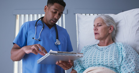 Young male registered nurse reviewing health record with senior female patient. African-American RN looking over doctors notes with elderly woman resting in hospital bed