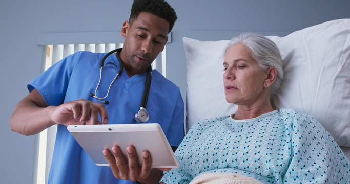 Wall Mural -  - Young african-american RN and senior patient looking at tablet computer at hospital. Male registered nurse showing ill elderly woman electronic pad