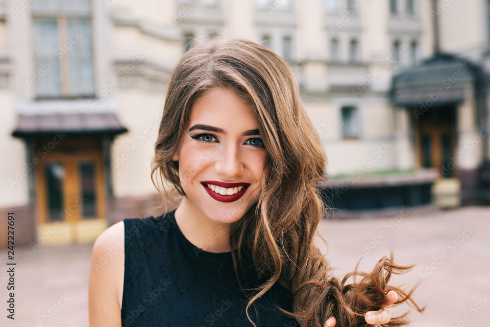 Closeup portrait of effective girl with long curly hair smiling to camera on street on building background. She wears black dress, vinous  lips. - obrazy, fototapety, plakaty 