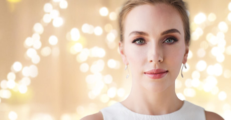 jewelry, luxury and people concept - portrait of woman in white dress with diamond earring over beige background and festive lights