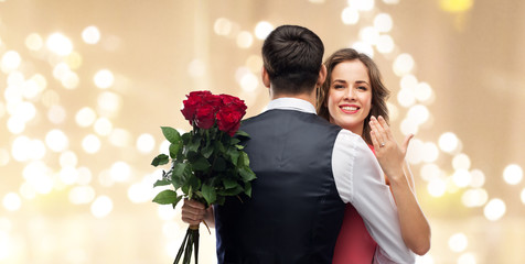 Poster - love, couple, proposal and people concept - happy woman with engagement ring and bunch of roses hugging man over beige background with festive lights