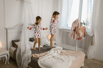 Wall Mural - Two little girls in their pajamas are having fun jumping on a bed in a sunlit cozy bedroom