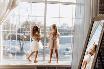 Wall Mural - Two little sisters dressed in the beautiful dresses stand on the windowsill next to the mirror