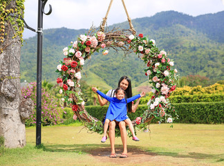 Asian mother and her daughter sitting on beautiful basket swing with the colorful roses flower in the nature garden hanging on pole under tree.