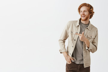 Studio shot of cool and famous handsome redhead male student with beard and wavy stylish hairstyle in beige jacket tilting and pointing left with finger gun gesture as if greeting mate on street