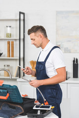 Wall Mural - profile of adult repairman holding pipe for kitchen faucet repairing and using smartphone