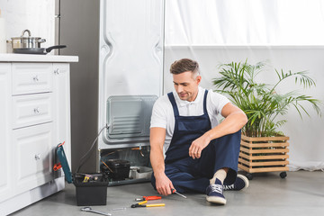 Wall Mural - tired adult repairman sitting on floor and looking at tools while repairing refrigerator