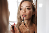 Young woman in lingerie underwear looking at mirror apply her lipstick lip gloss doing makeup.