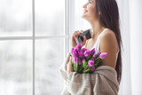 Fototapeta Tulipany - Young attractive woman talking on phone and smiling. Beautiful girl typing the message on the smartphone. Female at home indoors.
