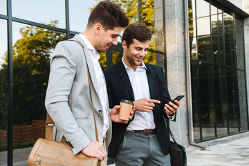 Wall Mural - Photo of young businessmen in suits using smartphones, while standing outdoor near building with takeaway coffee