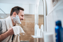 Portrait Of A Handsome Man With Shaving Cream On His Face And Towel While Standing In The Bathroom, Looking At Mirror.