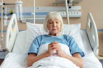 Wall Mural - senior woman lying in bed with folded hands and praying in hospital