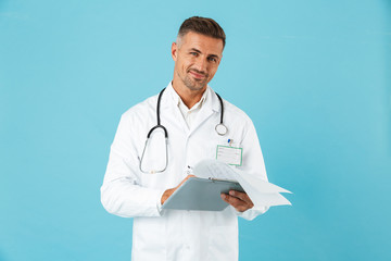 Canvas Print - Portrait of cheerful medical doctor with stethoscope holding health card, standing isolated over blue background