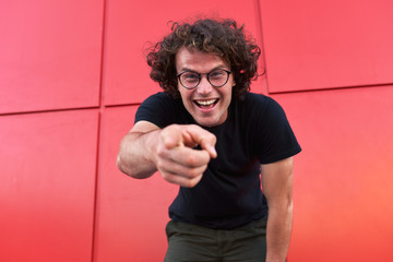 Poster - Image of happy handsome smiling male pointing with finger on the camera, posing for advertisement on red wall outdoor wearing black t-shirt and round eyeglasses. People emotions