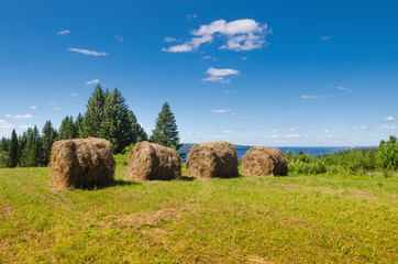 Wall Mural - beautiful summer landscape with sloping meadow, hay, forest and river with blue sky with clouds.