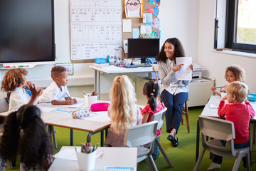 Smiling female infant school teacher sitting on a chair facing school kids in a classroom holding up and explaining a worksheet to them