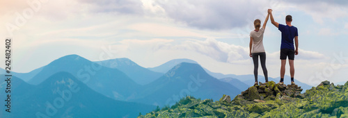Fotografía Back view of young tourist couple, athletic man and slim girl standing with raised arms holding hands on rocky mountain top enjoying fantastic panorama