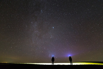 back view of two men with head flashlights on green grassy field under dark blue summer starry sky.