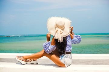 Wall Mural - Beautiful lady in a striped T-shirt and denim shorts, sunglasses and a straw hat. Positive emotions. Embankment on the background of the sea. The concept of summer holidays, relax, travel