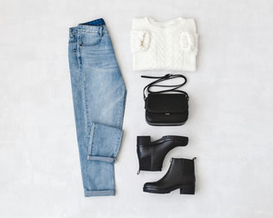 Blue jeans, white knitted sweater, small black cross body bag and leather ankle boots on grey background. Overhead view of woman's casual day outfits. Trendy hipster look. Flat lay.