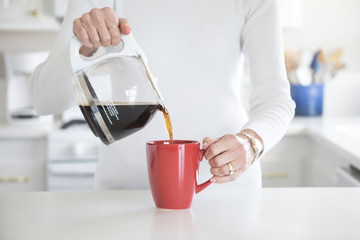 Photograph of a woman's hand pouring coffee into a red mug in a white kitchen