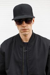 Portrait of a young handsome man hipster in a black cap in stylish black sunglasses in a fashionable autumn bomber jacket on a white background. American attractive guy.