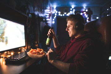 Wall Mural - Happy young man sits at night with a plate of chips and a bottle of beer in his hands, looks at the computer screen and smiles. Gamer plays video games on a computer and eats chips.