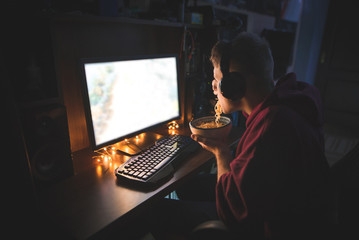 Wall Mural - Young man uses a computer at night at home and eats noodle soup. Gamer's back that plays video games on a computer and diner with fast food.