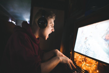 Expressive young gamer plays games at home coma in a cozy room at home. Portrait of an evil gamer in headphones playing video games on a computer.