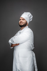 Poster - Side view of overweight bearded male in chef uniform keeping arms crossed and looking away while standing on gray background