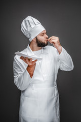 Poster - Handsome bearded male in chef uniform holding empty plate and kissing fingers while standing on gray background