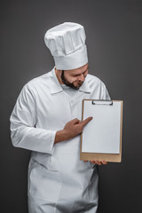 Poster - Bearded man in chef uniform pointing and looking at empty clipboard while standing on gray background and smiling