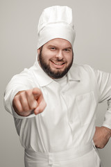 Sticker - Cheerful bearded man in chef uniform keeping hand on waist and pointing at camera while standing on light gray background