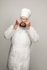 Poster - Overweight professional cook cheerfully smiling and holding pieces of ripe cucumber near eyes while standing on light gray background