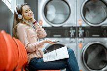 Young Woman Enjoying Music Waiting For The Clothes To Be Washed Sitting On The Chair At The Self-service Laundry
