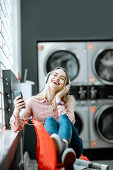 Wall Mural - Young woman enjoying music waiting for the clothes to be washed sitting on the chair at the self-service laundry