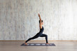 © Rithor - Young brunette woman fitness instructor wearing a black one-piece suit doing virabhadrasana yoga standing on a mat on a wooden floor against a background of a concrete wall