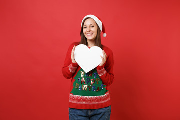 Smiling young Santa girl in knitted sweater, Christmas hat holding white blank empty heart isolated on bright red background. Happy New Year 2019 celebration holiday party concept. Mock up copy space.