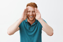 Studio Shot Of Playful And Joyful Good-looking Young Redhead Father With Bristle Holding Palms Along Face And Smiling Broadly, Playing Peekaboo With Kids And Having Fun In Family Circle Over Gray Wall