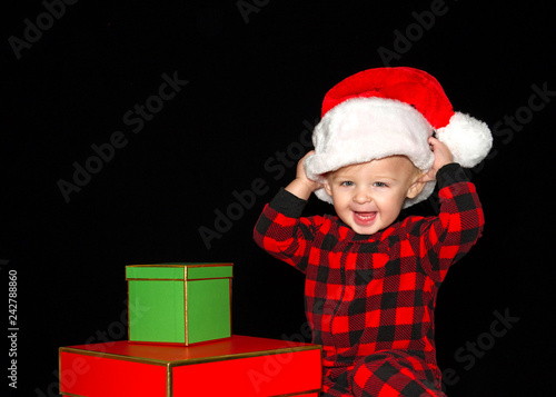 Fotografie, Obraz Close up portrait of one one year old baby boy wearing a Santa hat playing with red and green gift boxes, both hands on hat pulling onto head big smile with baby teeth showing