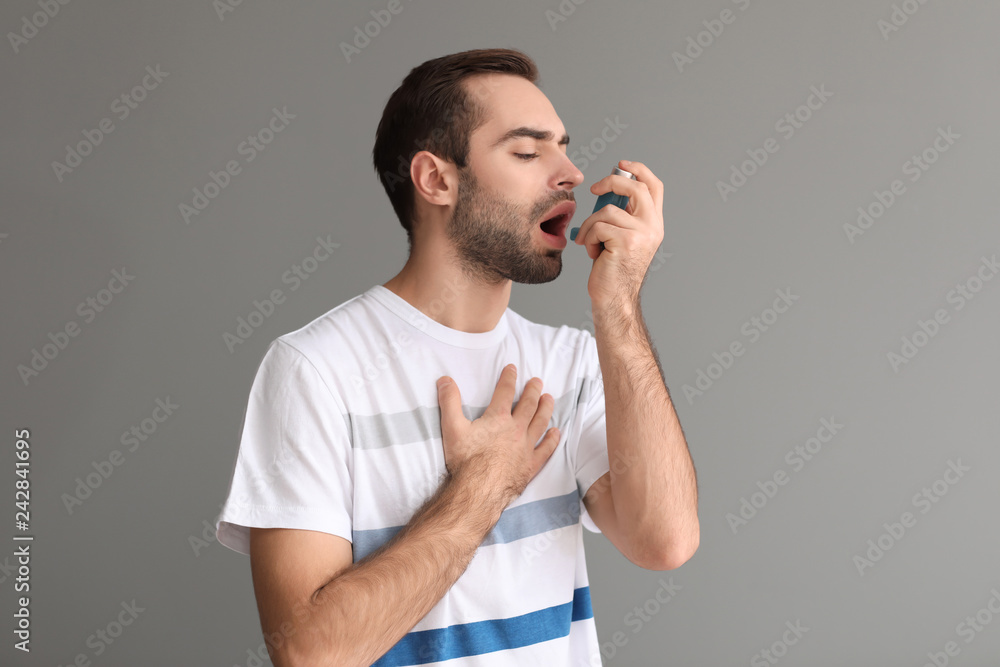 Young man with inhaler having asthma attack on grey background
