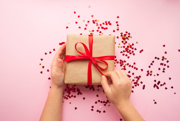 Wall Mural - Child is holding gift box wrapped in brown colored craft paper and tied with red bow on pink background with red heart shape confetti.