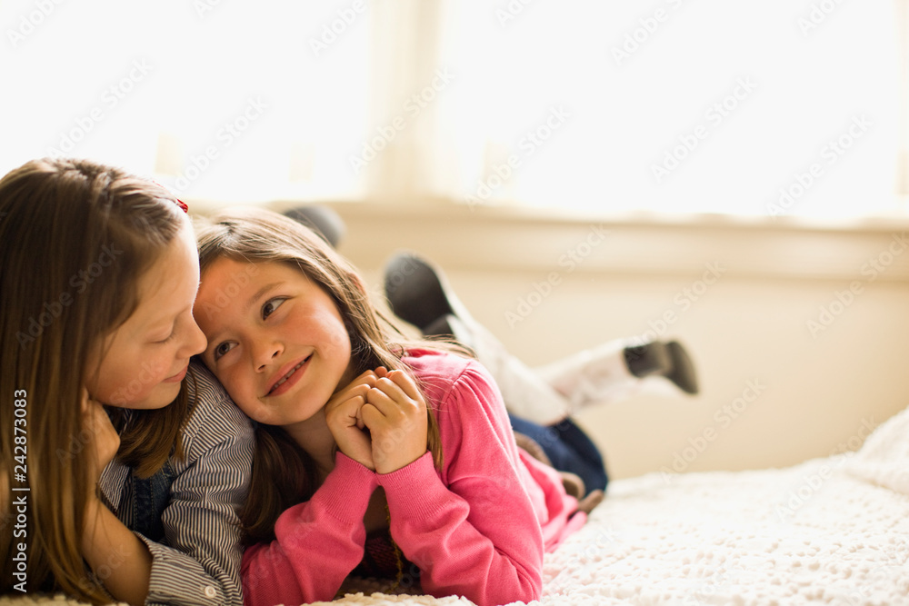 Two young girls smile at each other as they lie front down on a bed ...
