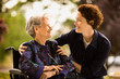 © Erickson Stock - Mature woman leans over to smile and talk to a senior woman with a oxygen nose hose (cannula) sitting in a wheelchair outside.