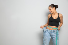 Slim Woman In Oversized Jeans With Measuring Tape On Light Background, Space For Text. Weight Loss