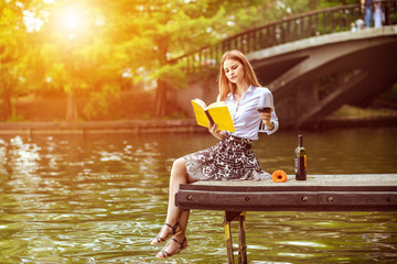 Young woman sitting on dock enjoying day in park reading book and drinking wine 