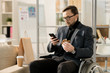 © Seventyfour - Serious businessman in eyeglasses sitting in wheelchair using his mobile phone while eating sandwich