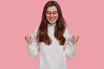 Wall Mural - Pretty young woman with tender smile points down with both index fingers, demonstrates new shoes she bought, wears white clothes, isolated over pink background. Design and advertising concept