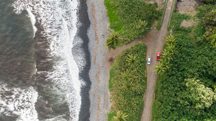 Stunning aerial drone view of a section of the famous Hana Highway south of Hana on the eastern side of the island of Maui, Hawaii. Beautiful black sand beach, waves, rainforest and two cars. 