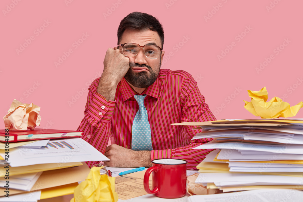 Lazy displeased fatigue male worker keeps fist on cheek, has sleepy look, dressed in formal shirt and tie, works with papers, has mess on workpace, poses over pink background, being overload - obrazy, fototapety, plakaty 